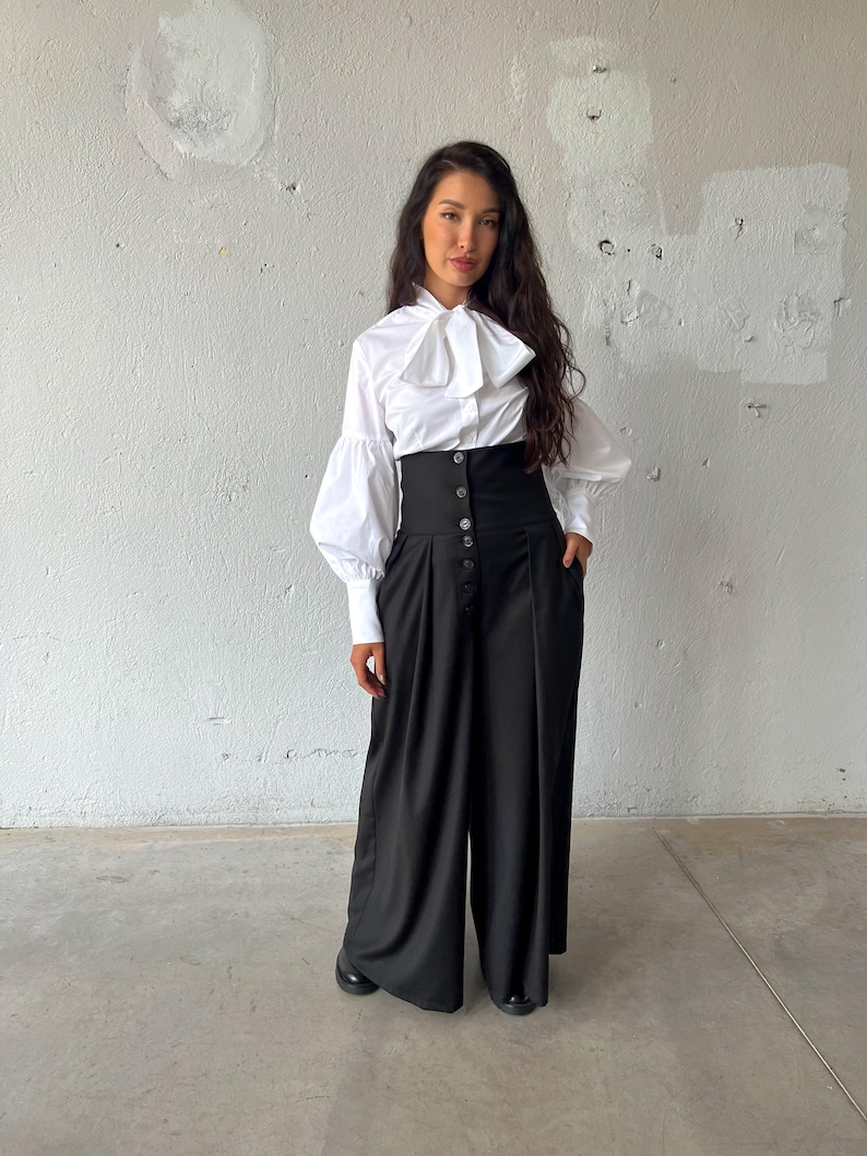 May include: A white blouse with a large bow and long, puffy sleeves paired with high-waisted black pants. The pants have a button-up closure and wide legs. The outfit is a stylish ensemble, suitable for various occasions.