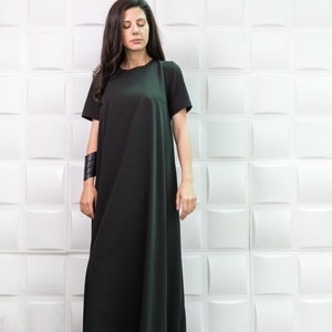 K&ouml;nnte beinhalten: Eine Frau tr&auml;gt ein langes, schwarzes Maxikleid mit kurzen &Auml;rmeln. Das Kleid hat einen schlichten, lockeren Schnitt und reicht bis zu den Kn&ouml;cheln. Sie steht vor einer wei&szlig;en Wand mit einem strukturierten Muster.