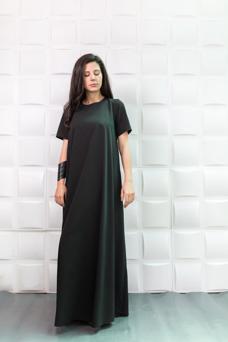 K&ouml;nnte beinhalten: Eine Frau tr&auml;gt ein langes, schwarzes Maxikleid mit kurzen &Auml;rmeln. Das Kleid hat einen schlichten, lockeren Schnitt und reicht bis zu den Kn&ouml;cheln. Sie steht vor einer wei&szlig;en Wand mit einem strukturierten Muster.