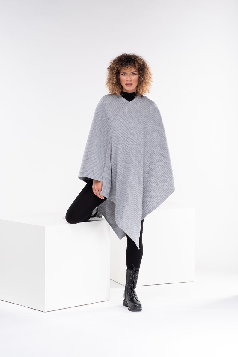 May include: A grey knitted poncho with a diagonal design. The poncho is being worn by a person with curly hair and black leggings. The person is standing on a white platform.