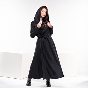 May include: A woman wearing a long black hooded coat with a belt. The coat has a flowing design and the woman is standing on a white background.