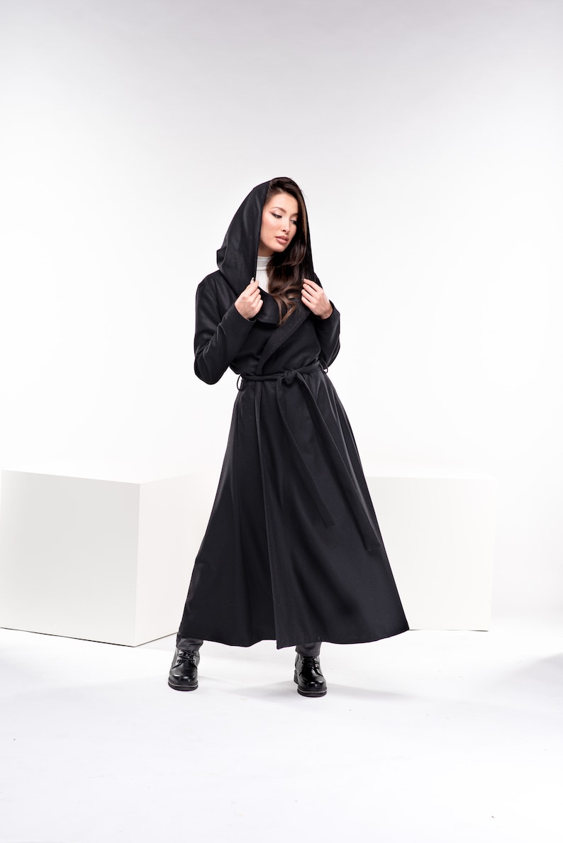 May include: A woman wearing a long black hooded coat with a belt. The coat has a flowing design and the woman is standing on a white background.