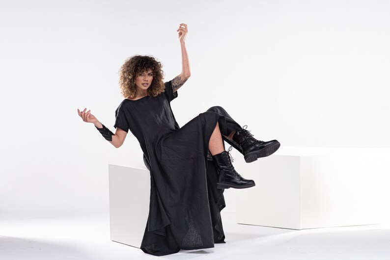 May include: A woman wearing a black linen dress and black boots sits on a white cube. She has curly brown hair and is wearing a black bracelet on her left wrist.