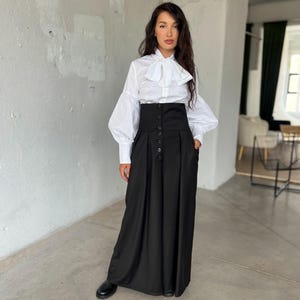 May include: A woman wearing a white blouse with a large bow and long sleeves, paired with a high-waisted black skirt. The skirt features a button-down front and pockets. Black shoes complete the outfit, creating a classic, elegant look.