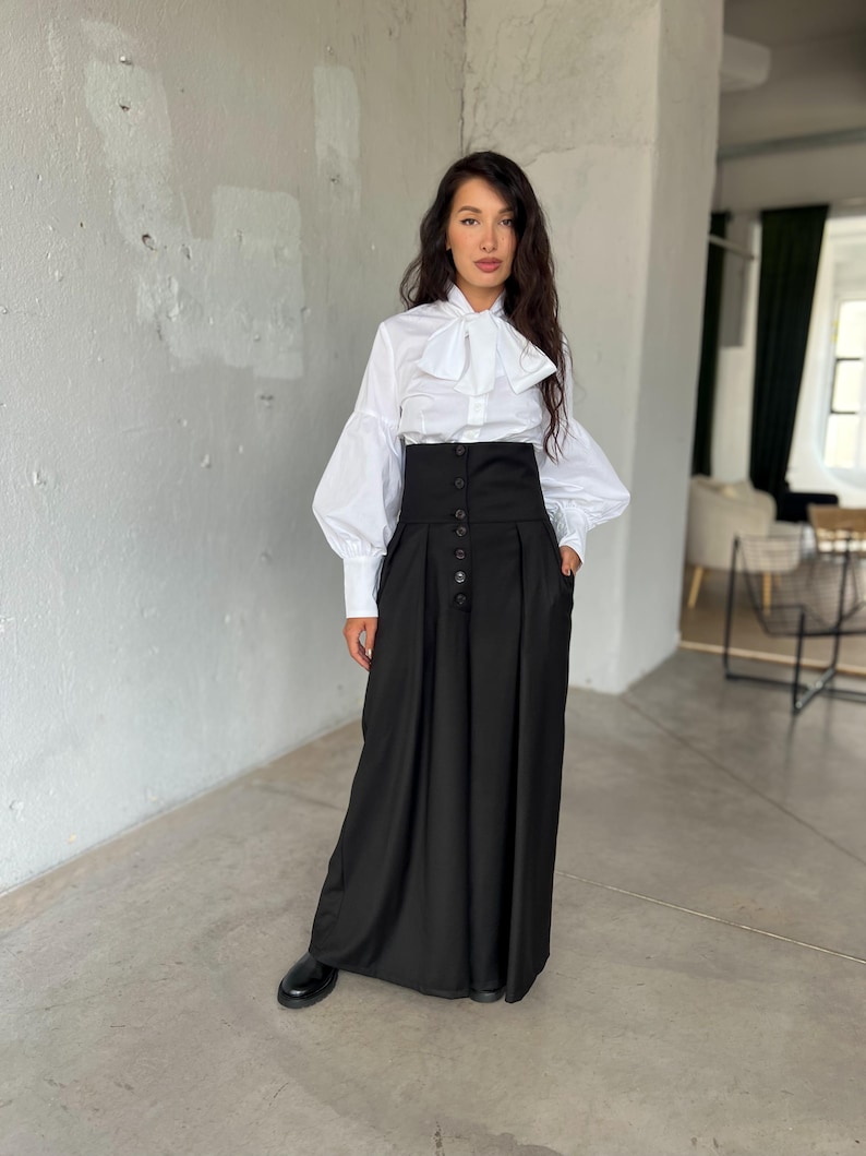 May include: A woman wearing a white blouse with a large bow and long sleeves, paired with a high-waisted black skirt. The skirt features a button-down front and pockets. Black shoes complete the outfit, creating a classic, elegant look.