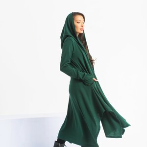 May include: A long, dark green hooded cardigan with a flowing, asymmetrical hem. The cardigan is made of a soft, knit fabric and has a relaxed fit.