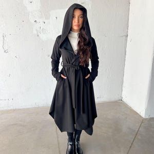 May include: A black hooded coat with a wrap-around design and a matching belt. The coat features long sleeves and pockets. The model is wearing black boots and a white turtleneck underneath the coat.