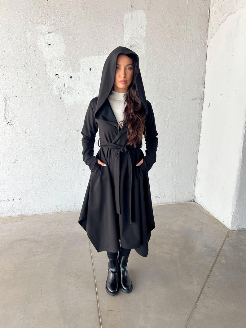 May include: A black hooded coat with a wrap-around design and a matching belt. The coat features long sleeves and pockets. The model is wearing black boots and a white turtleneck underneath the coat.