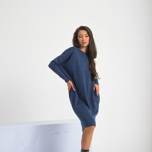 May include: A woman wearing a long, navy blue, loose-fitting sweater dress with long sleeves. She is wearing black lace-up boots and standing on a white surface.