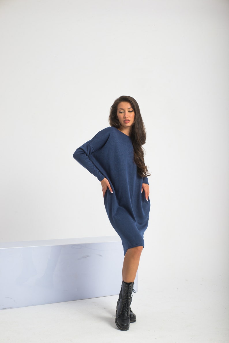 May include: A woman wearing a long, navy blue, loose-fitting sweater dress with long sleeves. She is wearing black lace-up boots and standing on a white surface.