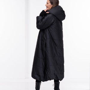 May include: A long black puffer coat with a hood. The coat has a quilted design and is being worn by a person standing in a white room.