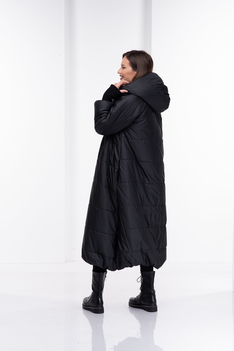 May include: A long black puffer coat with a hood. The coat has a quilted design and is being worn by a person standing in a white room.