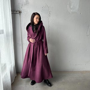 May include: A long, hooded coat in a deep burgundy color. The coat features a wrap-around design with a matching belt, and a full skirt. The model is wearing black boots. The coat is made of a soft, flowing fabric.