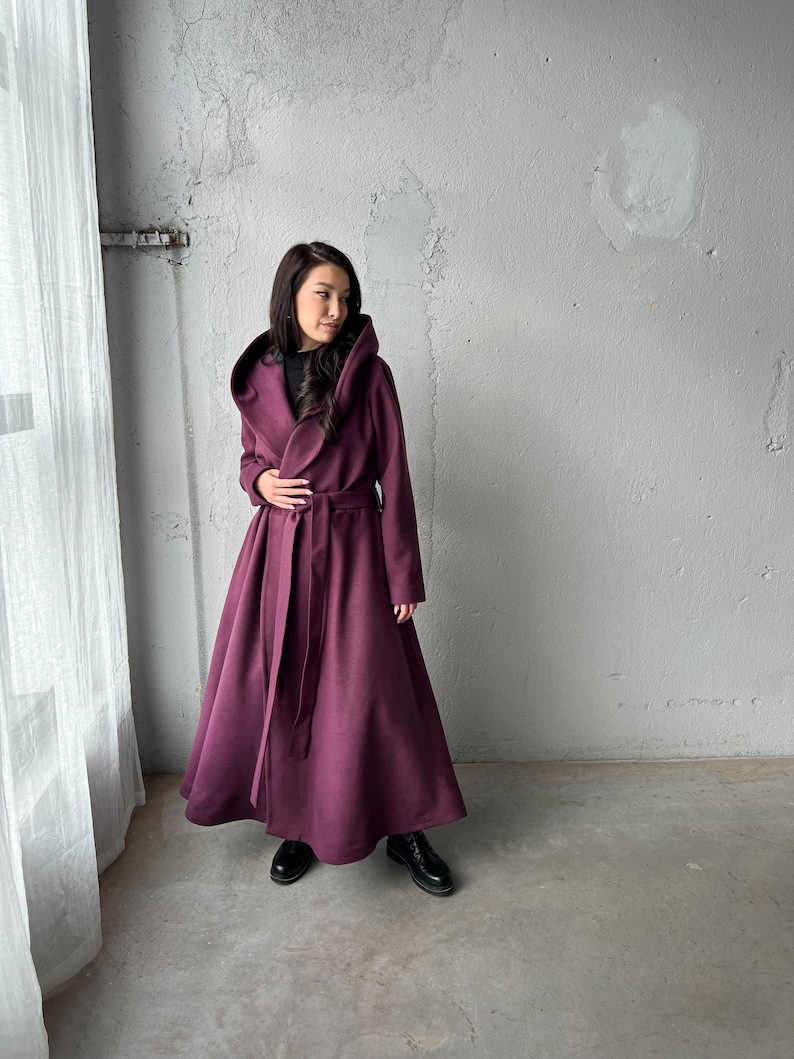 May include: A long, hooded coat in a deep burgundy color. The coat features a wrap-around design with a matching belt, and a full skirt. The model is wearing black boots. The coat is made of a soft, flowing fabric.