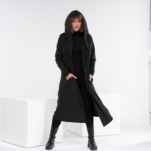 Pu&ograve; includere: Un lungo cardigan nero con cappuccio e tasche. La modella indossa pantaloni neri e stivali neri.