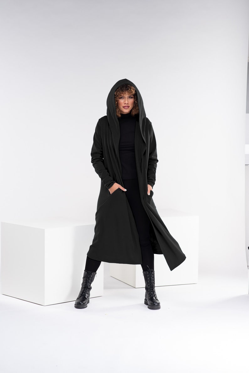 Pu&ograve; includere: Un lungo cardigan nero con cappuccio e tasche. La modella indossa pantaloni neri e stivali neri.