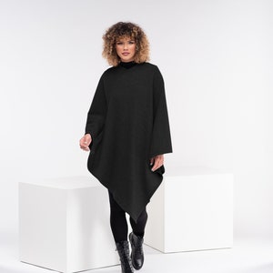 May include: A woman wearing a black knitted poncho with a long, asymmetrical hemline. She is standing on a white platform and wearing black leggings and black boots.