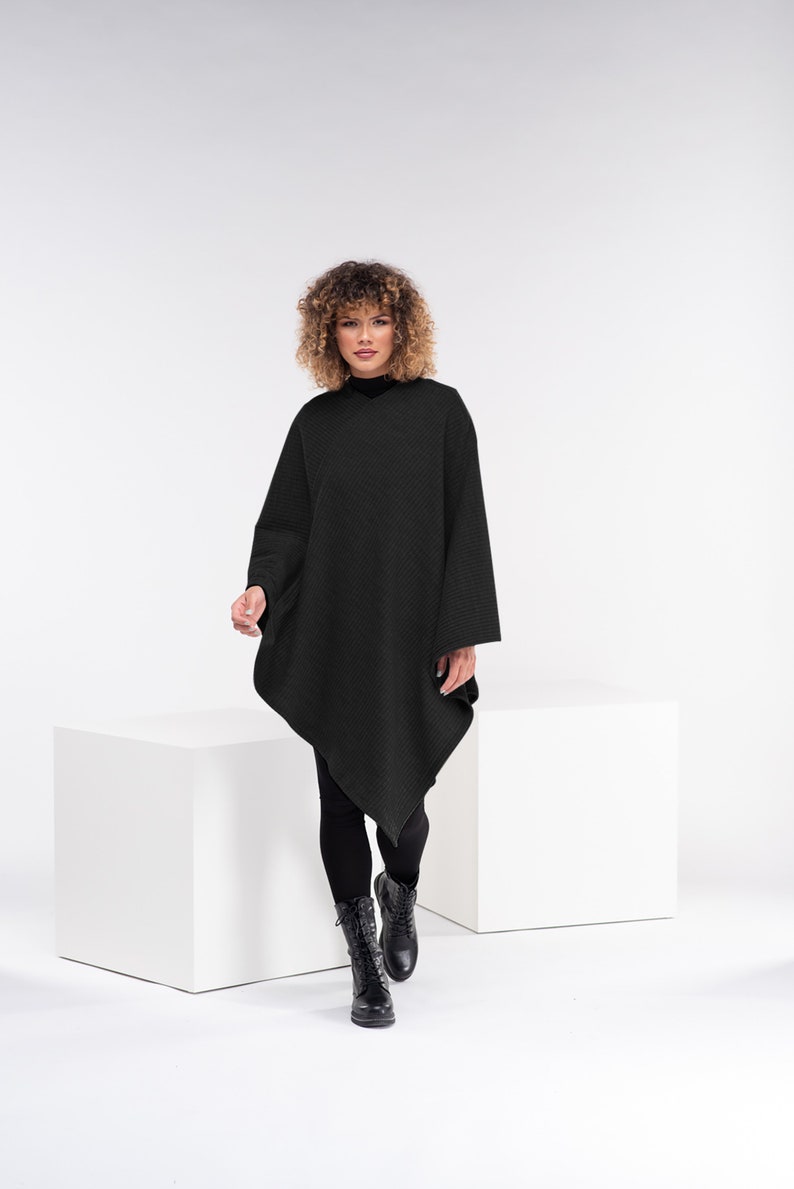 May include: A woman wearing a black knitted poncho with a long, asymmetrical hemline. She is standing on a white platform and wearing black leggings and black boots.