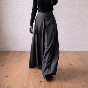 Op de afbeelding: Een broek met hoge taille en wijde pijpen die tot op de grond valt in een effen antracietgrijze kleur. De broek heeft opvallende verticale plooien die vanaf de tailleband naar beneden lopen, wat zorgt voor een volumineus, vloeiend silhouet.