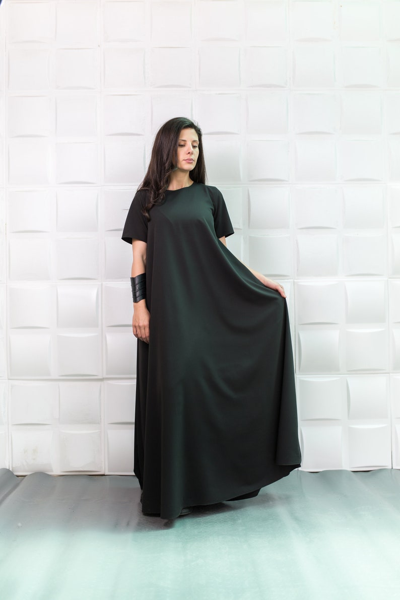 K&ouml;nnte beinhalten: Eine Frau tr&auml;gt ein langes, schwarzes, locker sitzendes Kleid mit kurzen &Auml;rmeln. Sie steht vor einer wei&szlig;en Wand mit einem strukturierten Muster.