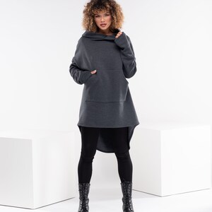 May include: A woman wearing a charcoal gray hooded sweatshirt with a long, asymmetrical hemline and a large front pocket. She is wearing black leggings and black lace-up boots.