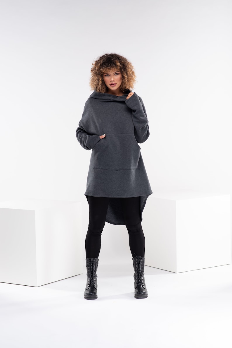 May include: A woman wearing a charcoal gray hooded sweatshirt with a long, asymmetrical hemline and a large front pocket. She is wearing black leggings and black lace-up boots.