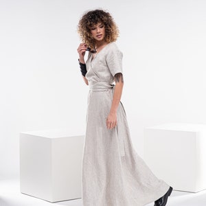 May include: A woman wearing a light beige linen wrap dress with a long skirt and short sleeves. She is standing in front of a white background and holding a pair of sunglasses.