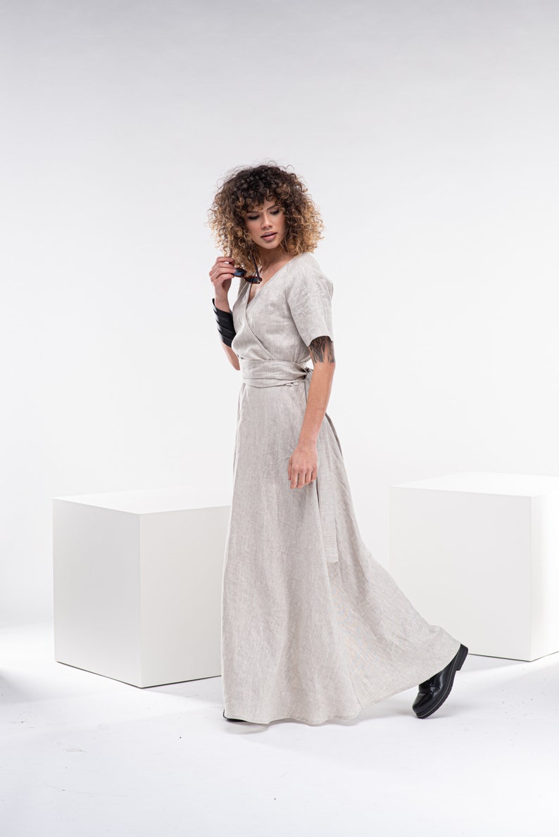 May include: A woman wearing a light beige linen wrap dress with a long skirt and short sleeves. She is standing in front of a white background and holding a pair of sunglasses.