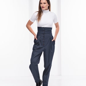 Pu&ograve; includere: Una donna indossa un top bianco a maniche corte e collo alto e pantaloni blu navy a vita alta. I pantaloni hanno una chiusura con bottoni e tasche. Stivali in pelle nera completano l'outfit. Lo sfondo &egrave; bianco.