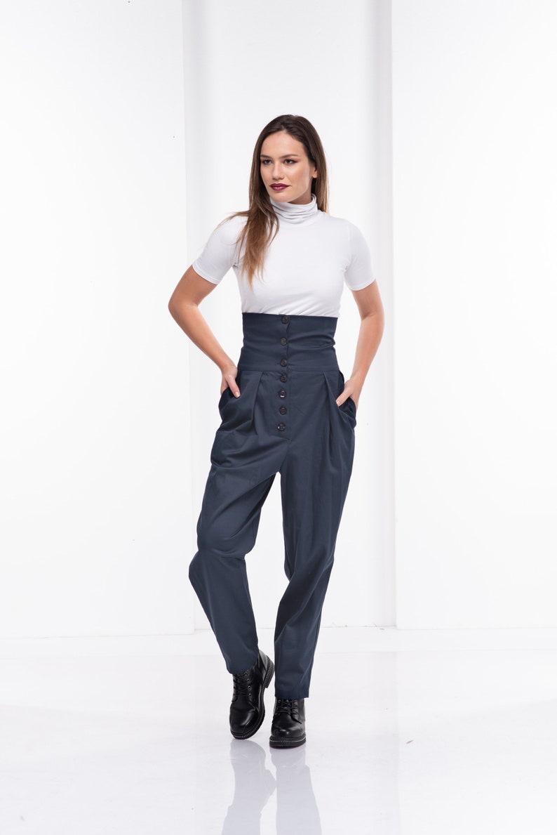 Pu&ograve; includere: Una donna indossa un top bianco a maniche corte e collo alto e pantaloni blu navy a vita alta. I pantaloni hanno una chiusura con bottoni e tasche. Stivali in pelle nera completano l'outfit. Lo sfondo &egrave; bianco.