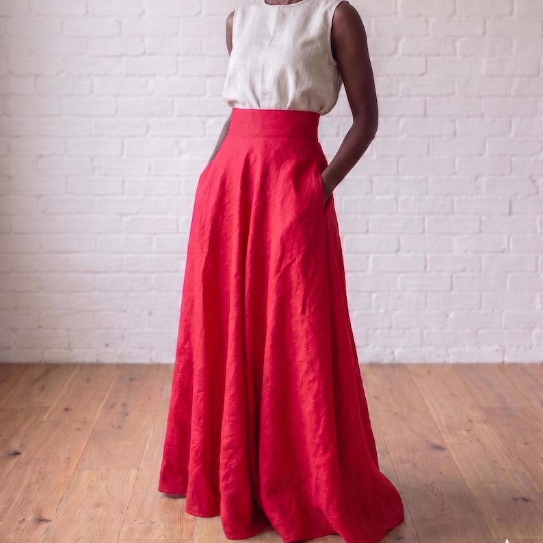 May include: A sleeveless cream-colored top paired with a high-waisted, floor-length red skirt. The skirt features a wide waistband, a full, flowing silhouette, and side pockets. The ensemble is constructed from a textured fabric with visible weave.