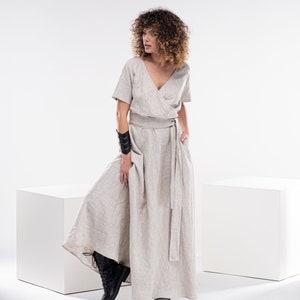 May include: A woman wearing a light beige linen dress with a wrap style bodice and a long skirt. She is wearing a black leather bracelet on her left wrist and black boots.