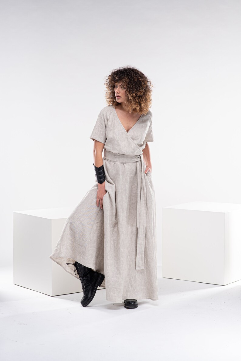 May include: A woman wearing a light beige linen dress with a wrap style bodice and a long skirt. She is wearing a black leather bracelet on her left wrist and black boots.