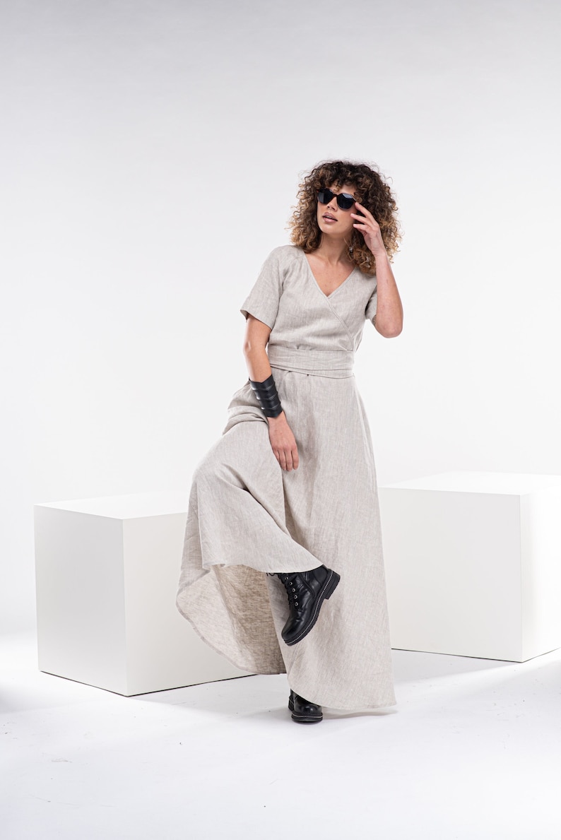 May include: A woman wearing a light beige linen dress with a V-neck and short sleeves. She is wearing black sunglasses and a black leather bracelet on her wrist. She is standing on a white platform with her leg bent and her foot resting on the platform.