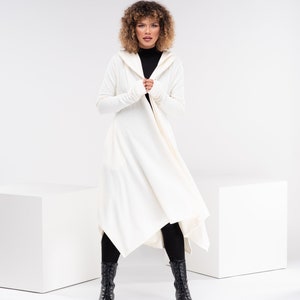 May include: A white hooded cardigan with long sleeves and a draped asymmetrical hem. The cardigan is worn over a black turtleneck and black leggings. The model is wearing black boots.