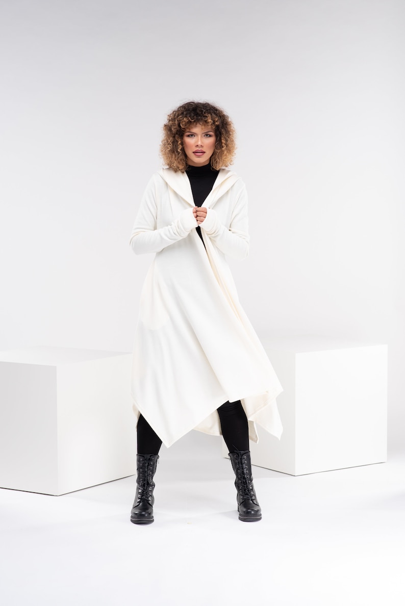 May include: A white hooded cardigan with long sleeves and a draped asymmetrical hem. The cardigan is worn over a black turtleneck and black leggings. The model is wearing black boots.