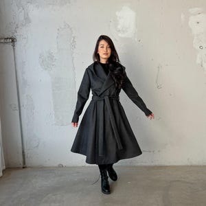 Puede incluir: Un abrigo largo de color gris oscuro con un dise&ntilde;o de envoltura y un cintur&oacute;n. El abrigo tiene una capucha y una falda acampanada.