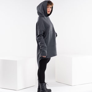 May include: A woman wearing a dark gray hooded sweatshirt with a long, asymmetrical hemline. She is wearing black leggings and black lace-up boots.
