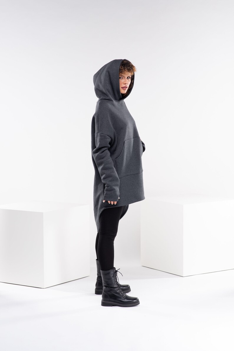 May include: A woman wearing a dark gray hooded sweatshirt with a long, asymmetrical hemline. She is wearing black leggings and black lace-up boots.