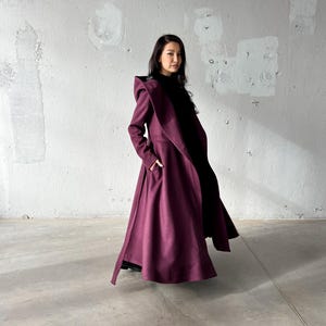 May include: A long, flowing, burgundy coat with a dramatic, oversized collar and lapel. The coat features a wide, flared skirt and pockets. The model is wearing a black top underneath. The coat is made of a heavy fabric, suitable for cooler weather.