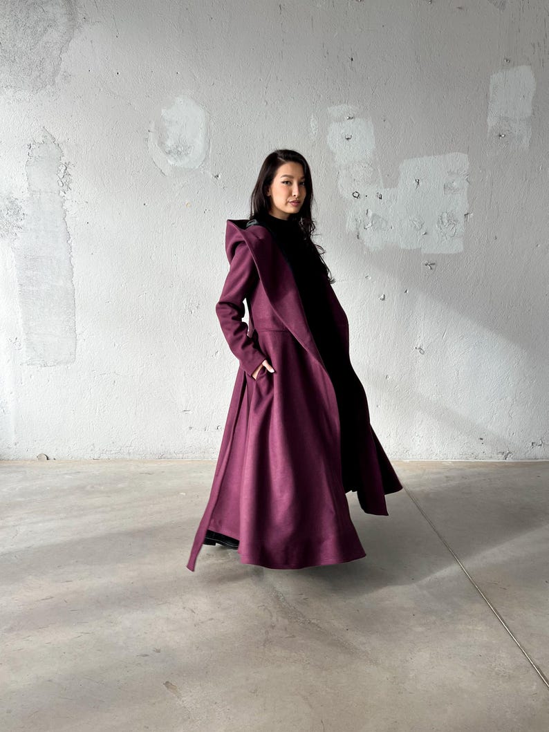 May include: A long, flowing, burgundy coat with a dramatic, oversized collar and lapel. The coat features a wide, flared skirt and pockets. The model is wearing a black top underneath. The coat is made of a heavy fabric, suitable for cooler weather.
