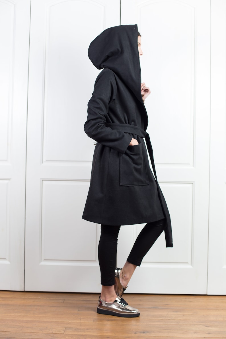 K&ouml;nnte beinhalten: Ein schwarzer Mantel mit Kapuze, passendem G&uuml;rtel und gro&szlig;en Taschen. Der Mantel ist knielang und wird mit schwarzen Leggings und silbernen Metallic-Schuhen getragen. Die Kapuze ist &uuml;bergro&szlig;.