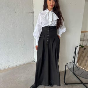 May include: A white blouse with a large bow and long sleeves paired with a high-waisted black skirt. The skirt features a button-down front and a long, flowing silhouette. The outfit is completed with black shoes.