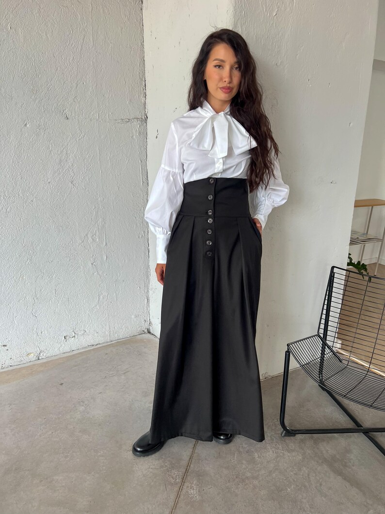 May include: A white blouse with a large bow and long sleeves paired with a high-waisted black skirt. The skirt features a button-down front and a long, flowing silhouette. The outfit is completed with black shoes.