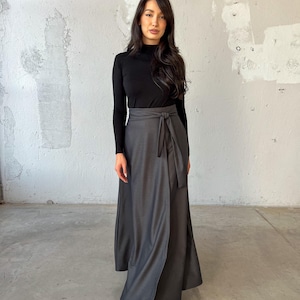 May include: A long, gray wrap skirt with a black tie belt. The skirt is made of a soft, flowing fabric and has a relaxed fit. The model is wearing a black turtleneck sweater.