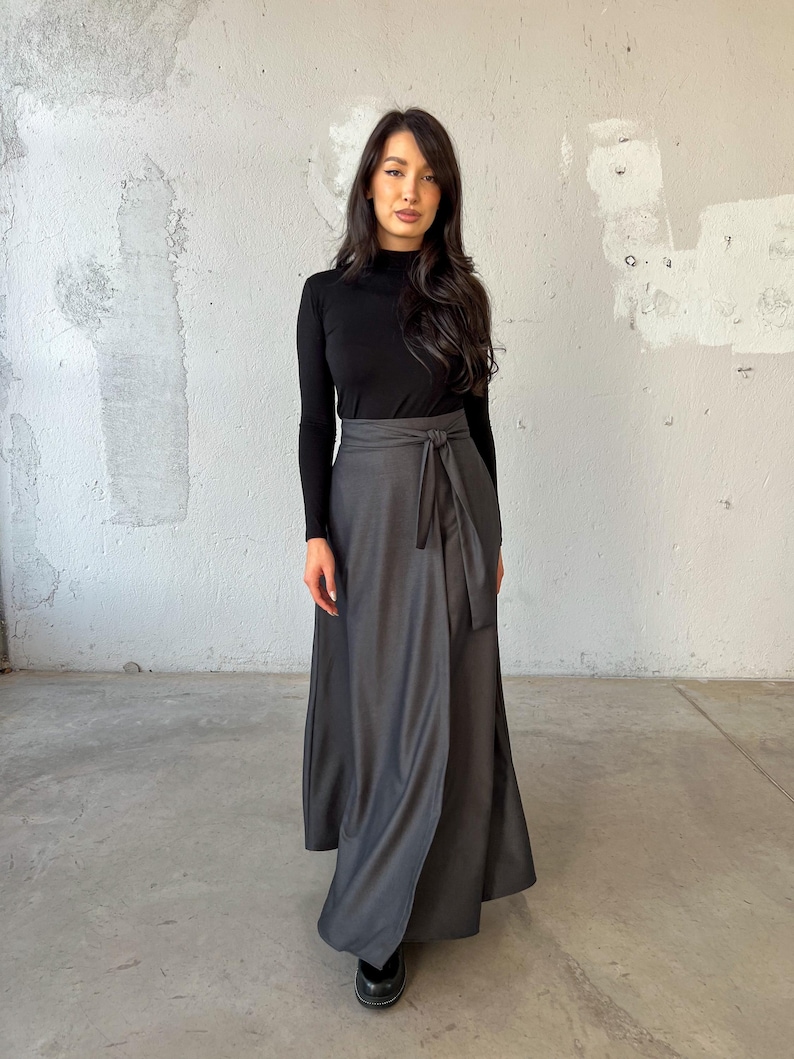 May include: A long, gray wrap skirt with a black tie belt. The skirt is made of a soft, flowing fabric and has a relaxed fit. The model is wearing a black turtleneck sweater.