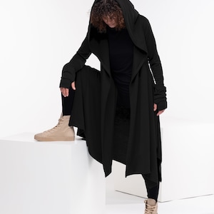May include: A black hooded long-sleeved cardigan with a long, flowing hemline. The cardigan is worn over a black long-sleeved shirt and black pants. The person is wearing beige high-top sneakers.