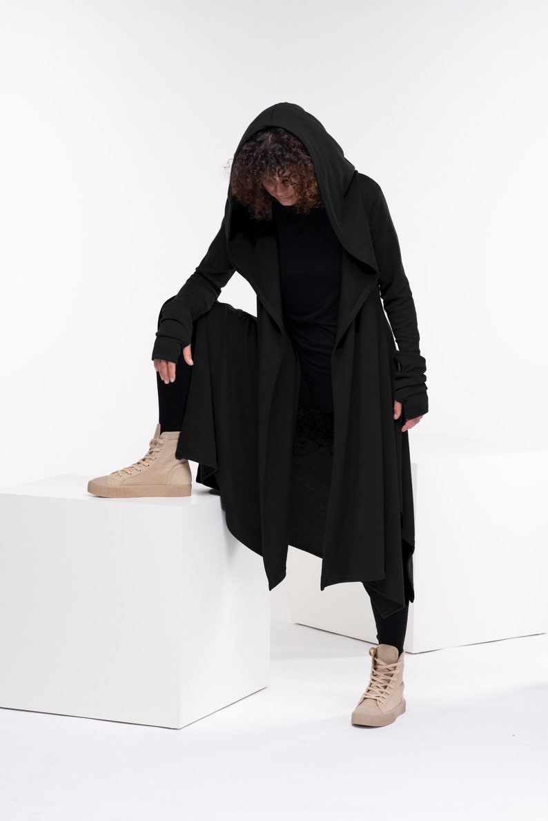 May include: A black hooded long-sleeved cardigan with a long, flowing hemline. The cardigan is worn over a black long-sleeved shirt and black pants. The person is wearing beige high-top sneakers.