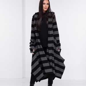 Op de afbeelding: Lange cardigan met capuchon in een zwart-grijze horizontale streep. De vloerlange cardigan wordt gedragen met een zwarte broek en laarzen. De mouwen zijn lang en de stijl is casual en modern.