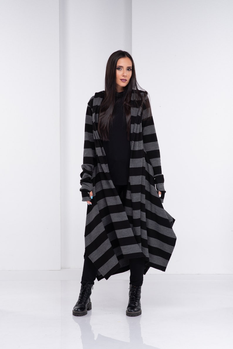 Op de afbeelding: Lange cardigan met capuchon in een zwart-grijze horizontale streep. De vloerlange cardigan wordt gedragen met een zwarte broek en laarzen. De mouwen zijn lang en de stijl is casual en modern.
